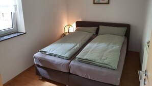 En liten, välordnad säng med ett enkelt nattduksbord och en lampa, ett fönster med utsikt och en inramad tavla på väggen.