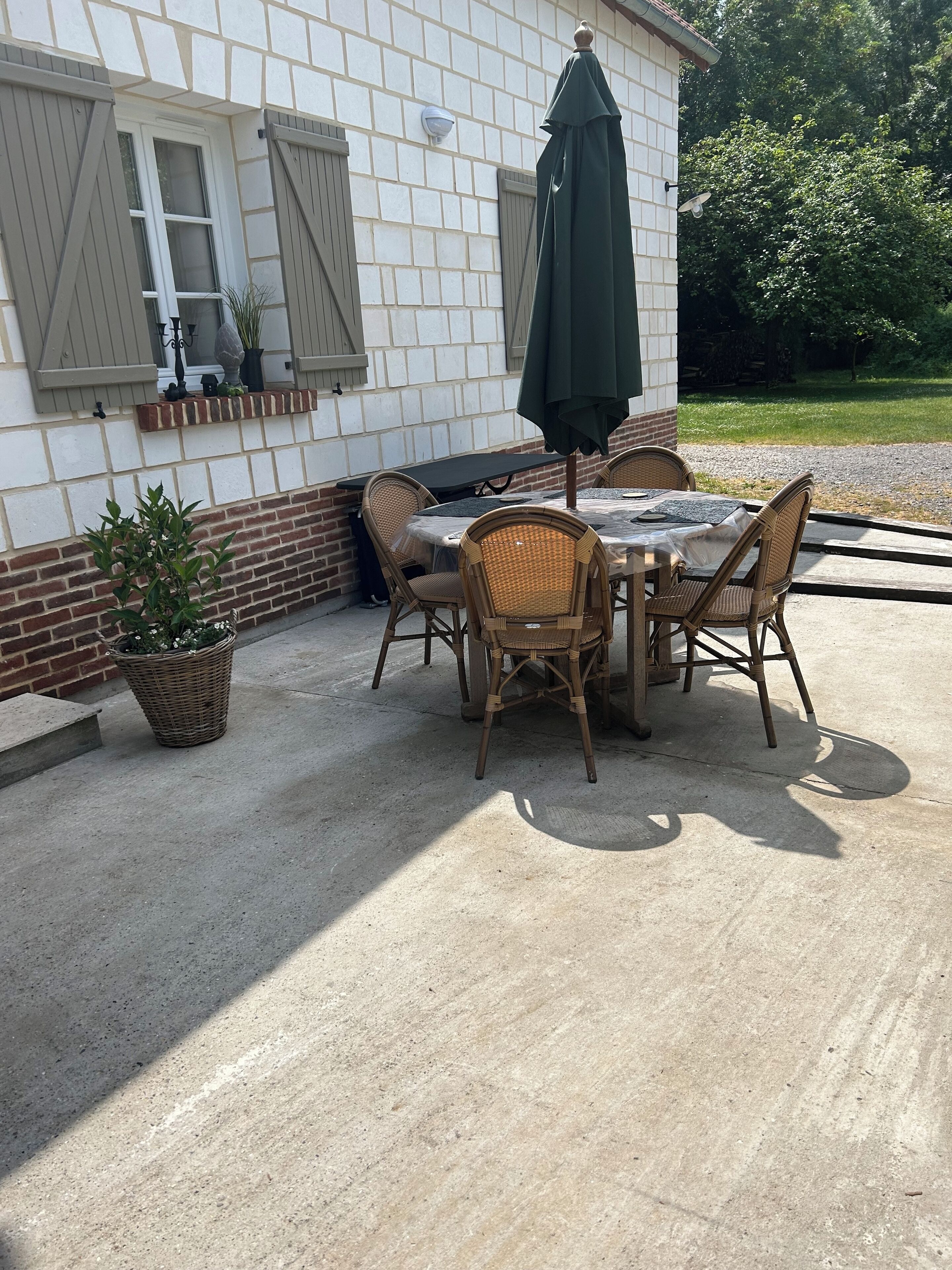 Een terras buiten met een tafel, stoelen, een groene parasol en een bloempot.