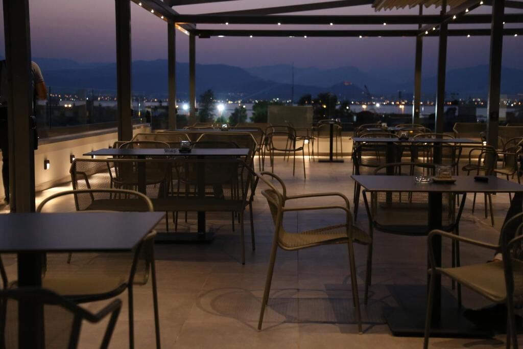 An outdoor dining area with tables and chairs, a pergola, and a view of the city and mountains at dusk.