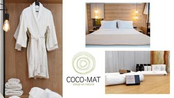 A hotel room with a wooden headboard, a bed with white linens, a bedside table with a lamp, and a wall-mounted robe rack with towels.
