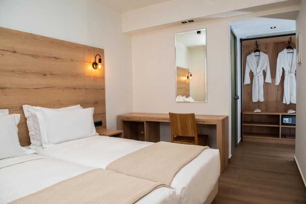 Habitación de hotel con cabecera de madera, un escritorio con silla, cama con ropa de cama blanca, un espejo y un vestidor con batas.