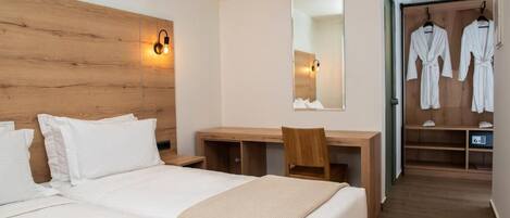 Habitación de hotel con cabecera de madera, un escritorio con silla, cama con ropa de cama blanca, un espejo y un vestidor con batas.