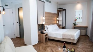 A modern hotel room with a wooden herringbone floor, a bed with a white duvet, bedside tables, a small wooden nightstand, a mirror, and a wall-mounted light fixture. - Liberty Hotel of Nafplio (Nafplio)
