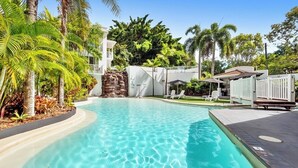 A swimming pool with clear water, surrounded by palm trees and a white building. - 1 Bedroom Apartment 301/1 in Alassio Resort (Palm Cove)