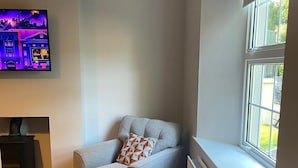 Een hoekappartement met een grijze bank, een kussen met een patroon, een flatscreen televisie aan de muur, een raam met uitzicht en een radiator onder het raam.