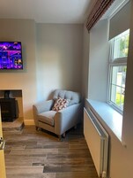 A corner room with a grey sofa, a patterned throw pillow, a flat-screen TV on the wall, a window with a view, and a radiator below the window.