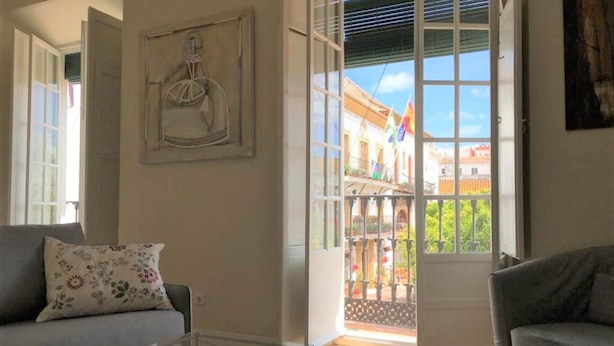 A spacious living room with a glass coffee table, a grey sofa, and a large window offering a view of a colorful building outside. - Cozy Studio in Marbella Old Town, Steps from Beach & Historic Sights (Marbella)