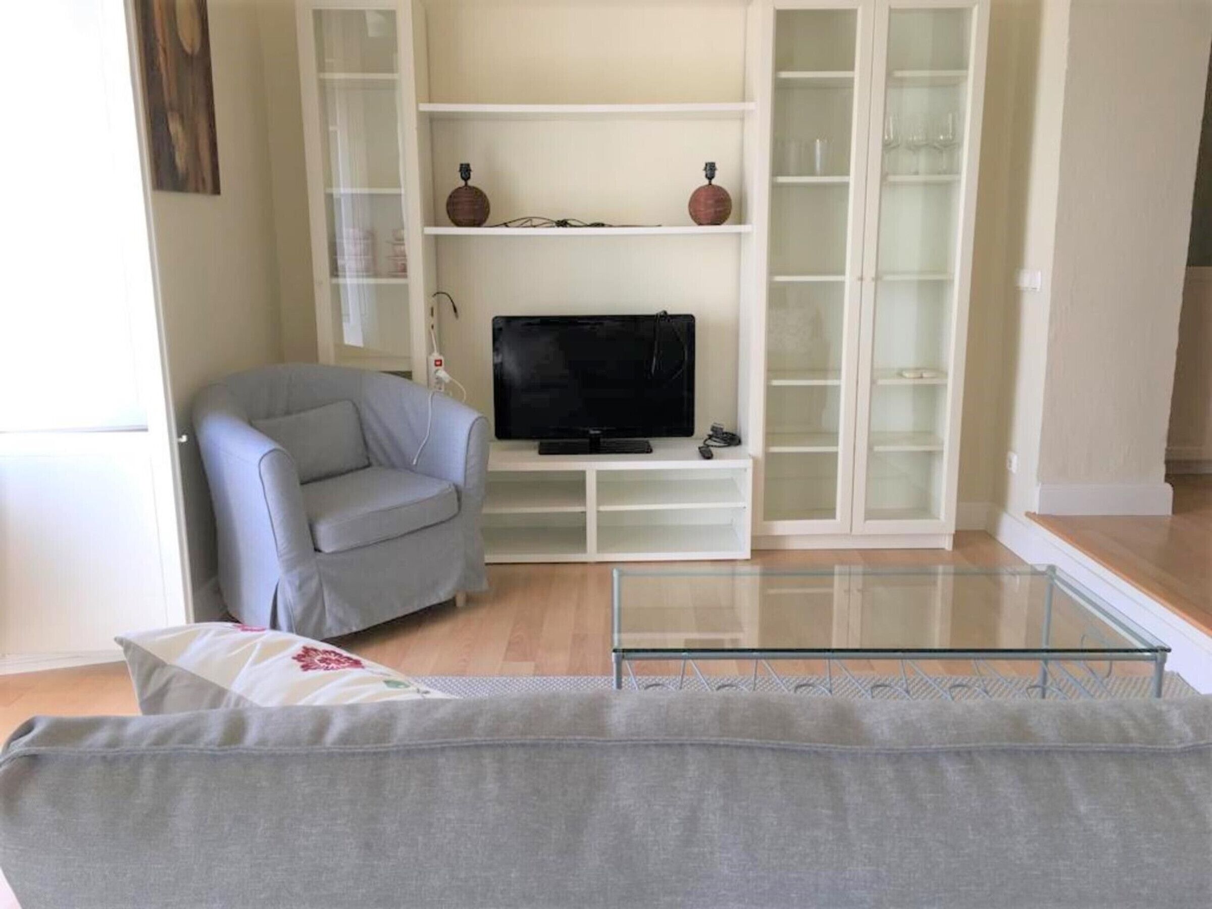 Una sala de estar con un sofá gris, un televisor sobre un mueble blanco y una mesa de centro de vidrio.