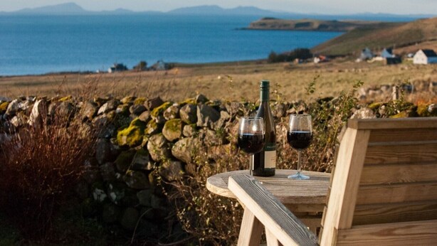 A wooden table with a bottle of wine and two glasses, set against a scenic coastal backdrop with stone walls and distant houses.