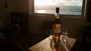 A bottle of Talisker Port Ruighle whisky on a wooden table with two glasses, in a room with a view of the sea through a window.