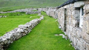 A stone wall pathway leading to a stone building with a window, surrounded by lush green grass and hills in the background.