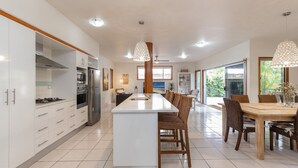 A modern kitchen with a central island, dining area with wicker chairs, and large windows offering a view. - Outrigger @ Boomerang Beach (Boomerang Beach)