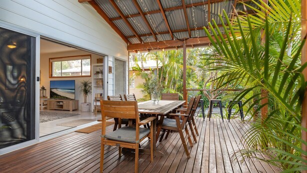 An outdoor patio with a wooden table and chairs, surrounded by lush greenery and a corrugated metal roof. - Outrigger @ Boomerang Beach (Boomerang Beach)