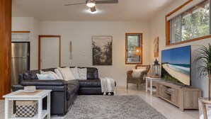 A living room with a dark leather sofa, a white coffee table, a grey shag rug, a wicker chair, a wooden cabinet, and a large beach painting. - Outrigger @ Boomerang Beach (Boomerang Beach)