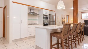 A modern kitchen with white cabinetry, a central island, bar stools, and a decorative light fixture. - Outrigger @ Boomerang Beach (Boomerang Beach)