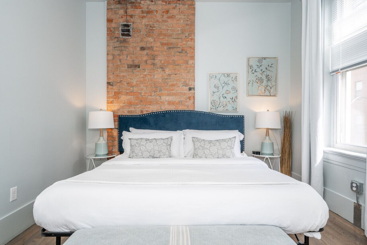 A bedroom with a brick wall, a bed with white bedding, two bedside tables with lamps, and a window with blinds.