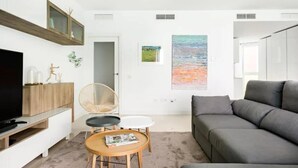 Un salon moderne avec un canapé gris, un téléviseur posé sur un meuble en bois, une table ronde en bois sur laquelle se trouve un magazine, un fauteuil en osier et un tableau accroché au mur.