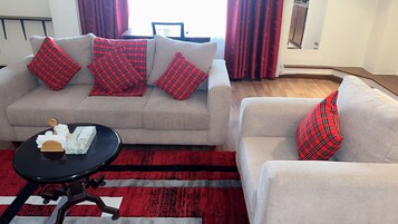 A living room with a grey sofa and a smaller armchair, both with red plaid cushions. A black coffee table with a white object and a wooden tray is in front of the sofa.