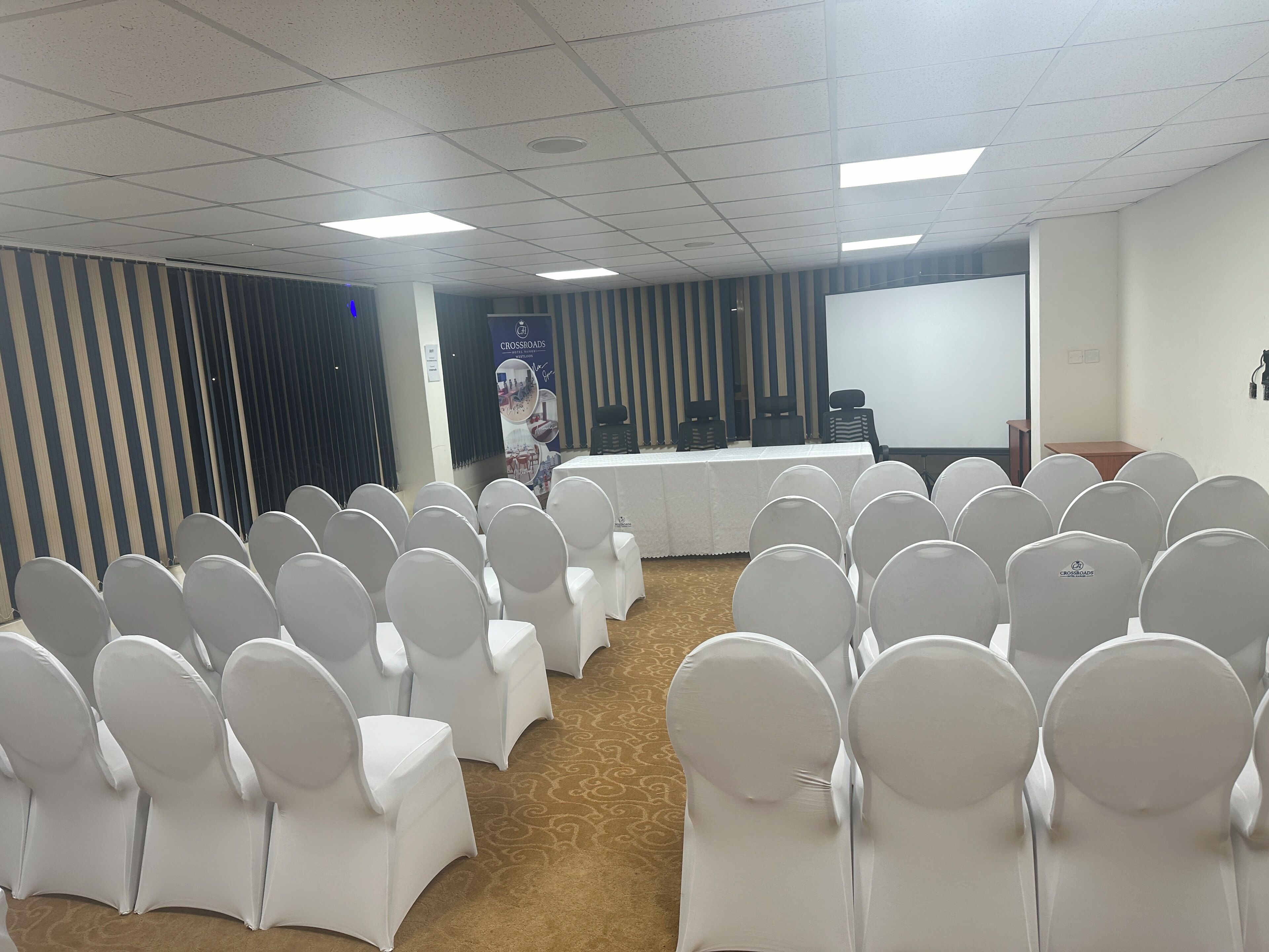 A conference room with rows of white chairs, a long table, and a projector screen.