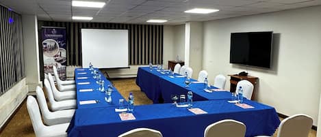 Un salón de conferencias con mesas largas, sillas blancas, una pantalla de proyector y un televisor en la pared.