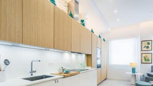 Una cucina moderna con mobili bianchi, un lavello in acciaio inox e un piano di lavoro in legno. Si vede una finestra con vista sull'esterno, e un piccolo tavolo rotondo con una lampada.