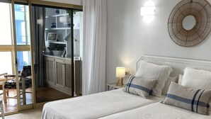 A bedroom with two beds, a nightstand with a lamp, a wall-mounted circular light, and a sliding glass door leading to a balcony.