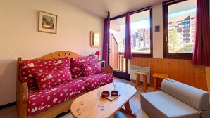 A cozy room with a wooden sofa, a small table with a glass, and a view of buildings through large windows.