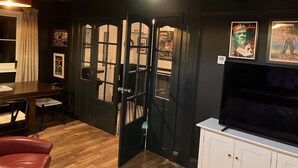 Ein Zimmer mit Holzboden, einem roten Sessel, einem Esstisch, einer schwarzen Tür mit Glasfeldern, einem weißen Schrank und einem Fernseher obenauf.