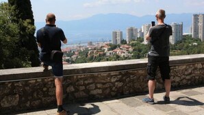 Deux personnes se tiennent sur une plateforme en pierre, d’où elles dominent une vue sur la ville, avec des bâtiments et des montagnes à l’horizon.