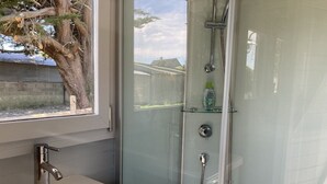 Ein Badezimmer mit einem weißen Waschbecken, einer Duschkabine aus Glas und einem Fenster mit Blick auf einen Garten.