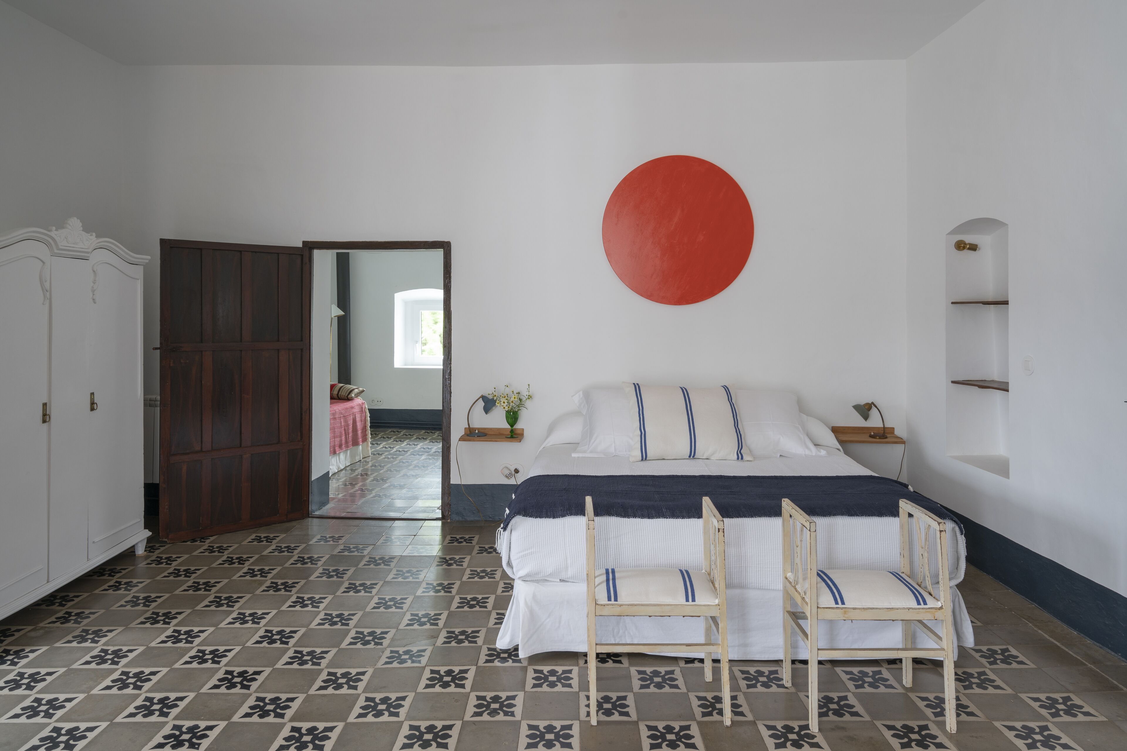 A bedroom with a bed, two chairs, a red circular decoration on the wall, a wardrobe, and a door leading to another room.