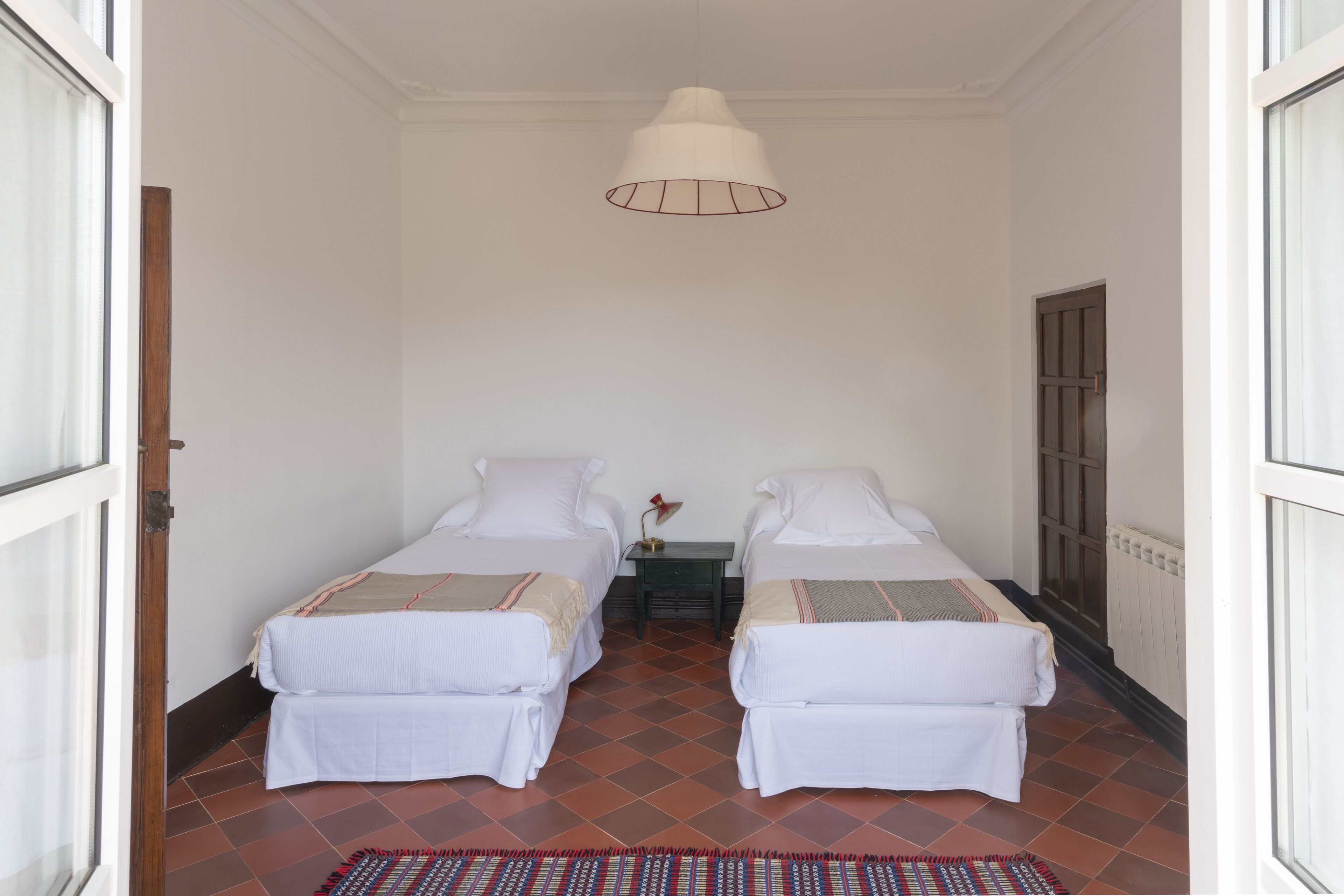 A room with two beds, a hanging light fixture, a small table with a lamp, and a patterned rug.