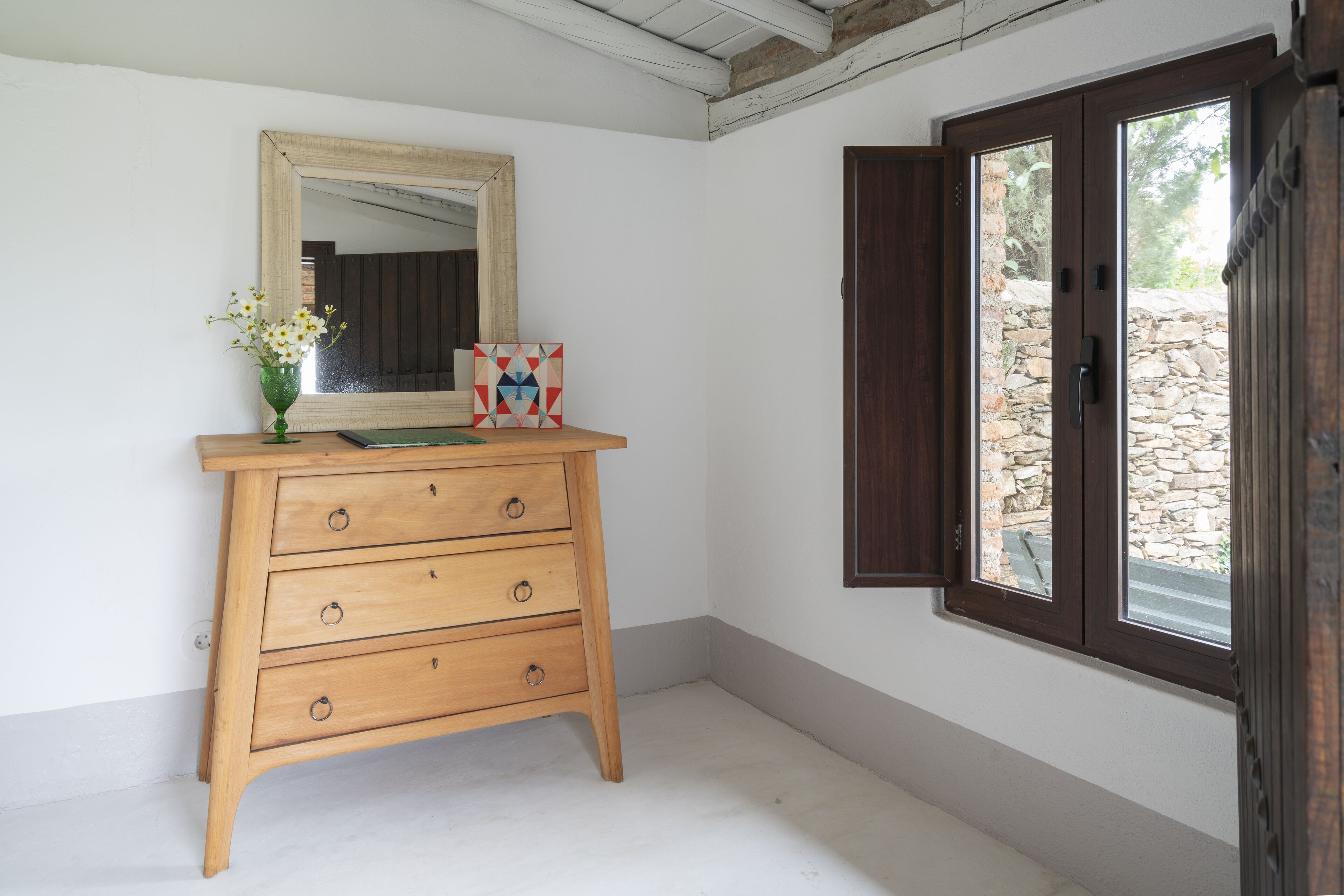 Een houten dressoir met spiegel en een bloevas, een geometrisch kunstwerk, en een raam met uitzicht op een stenen muur.