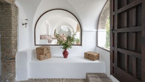 A white-walled room with a large arched mirror, a red vase with green plants, and wooden boxes on a bench.