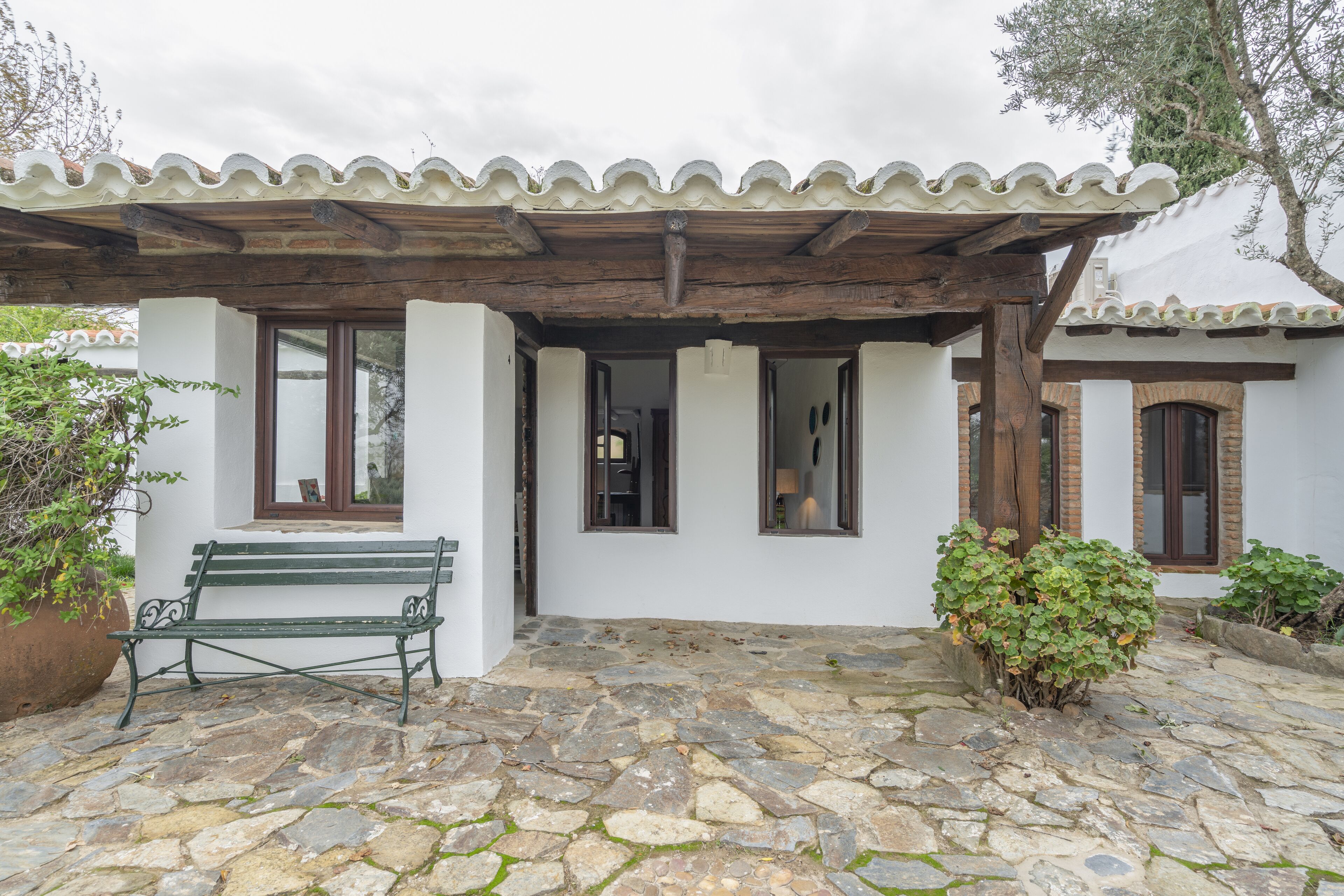 Une cour pavée de pierres, avec un banc en bois, un bâtiment blanc doté d’un toit en tuiles et un petit jardin.