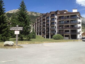 Um edifício de vários andares com varandas, uma placa indicando estacionamento privativo e montanhas ao fundo.