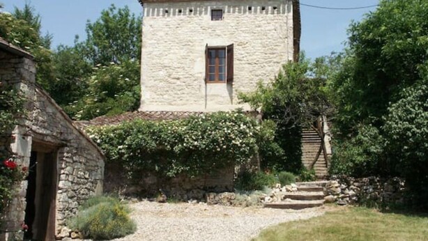 Ein Steinhaus mit einem kleinen Fenster und einer Tür, umgeben von Grünpflanzen und einem Kiesweg.