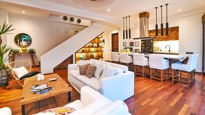 A modern living room with a staircase, white sofas, a wooden coffee table, and a kitchen area with bar stools. - Villa Kalita (Seminyak)
