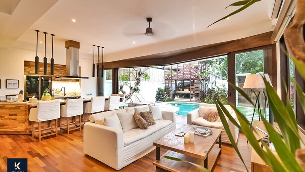 A modern living room with a white sofa, coffee table, and bar area with stools. There is a large window providing a view of the outdoor pool and garden. - Villa Kalita (Seminyak)