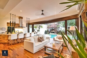 A modern living room with a white sofa, coffee table, and bar area with stools. There is a large window providing a view of the outdoor pool and garden. - Villa Kalita (Seminyak)