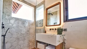 A bathroom with a hexagonal tile wall, a glass shower enclosure, a wooden floor, a white sink with a mirror, and a toilet. - Villa Kalita (Seminyak)
