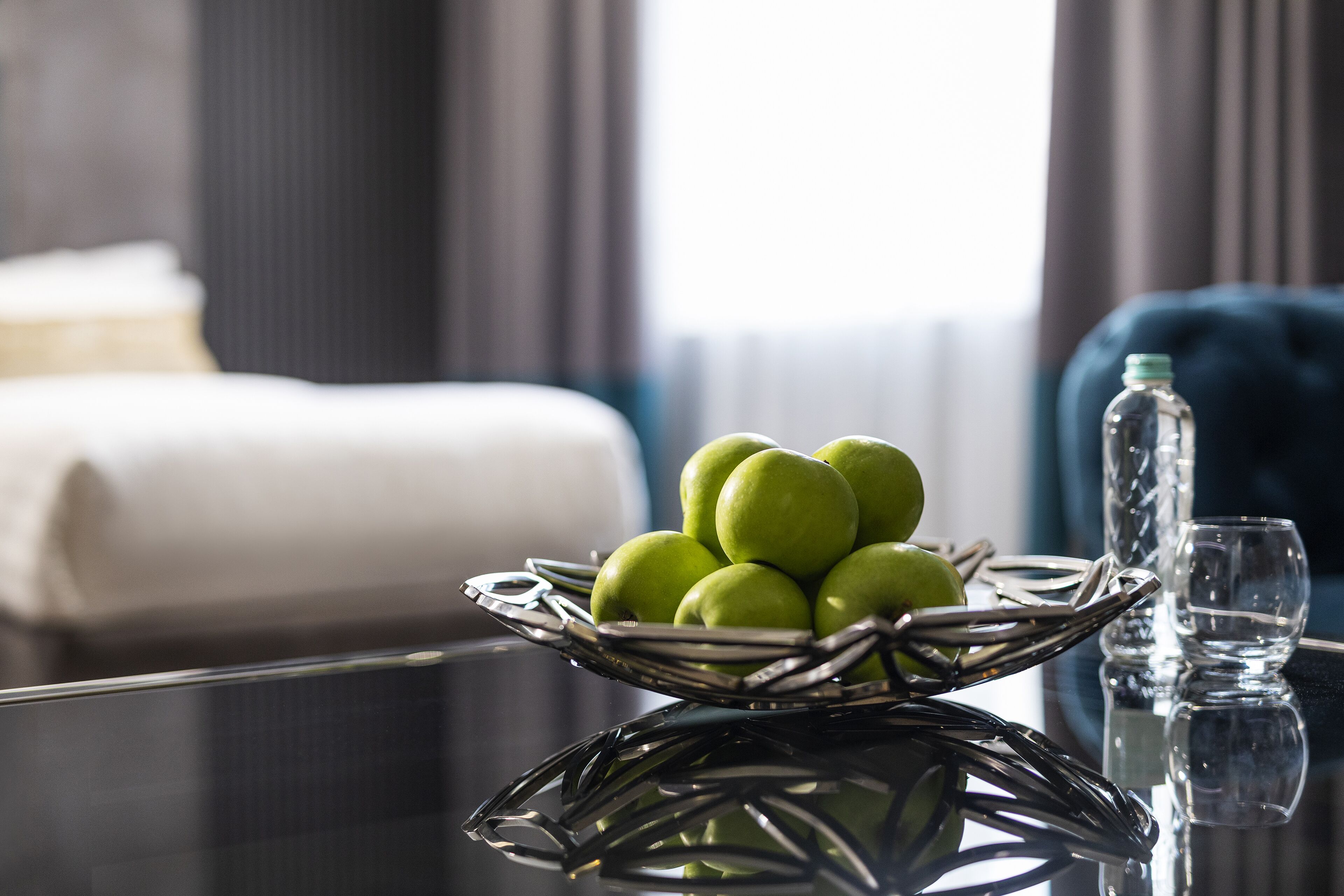A room with a glass table displaying a wicker bowl of green apples, a clear glass bottle, and a glass.