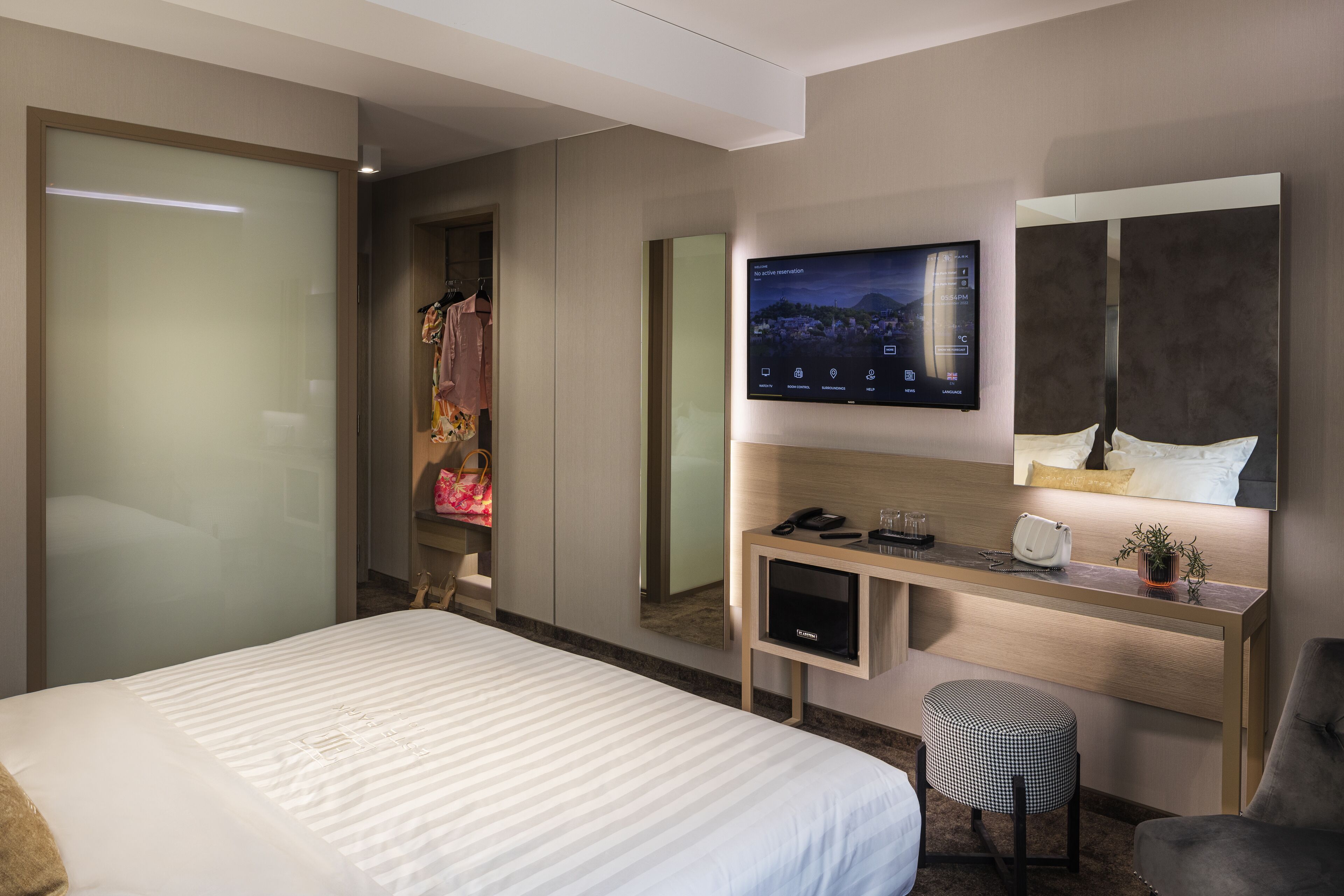 Ein modernes Hotelzimmer mit einem großen Bett, einem Schreibtisch, einem Flachbildfernseher, einem Spiegel und einem kleinen Hocker.