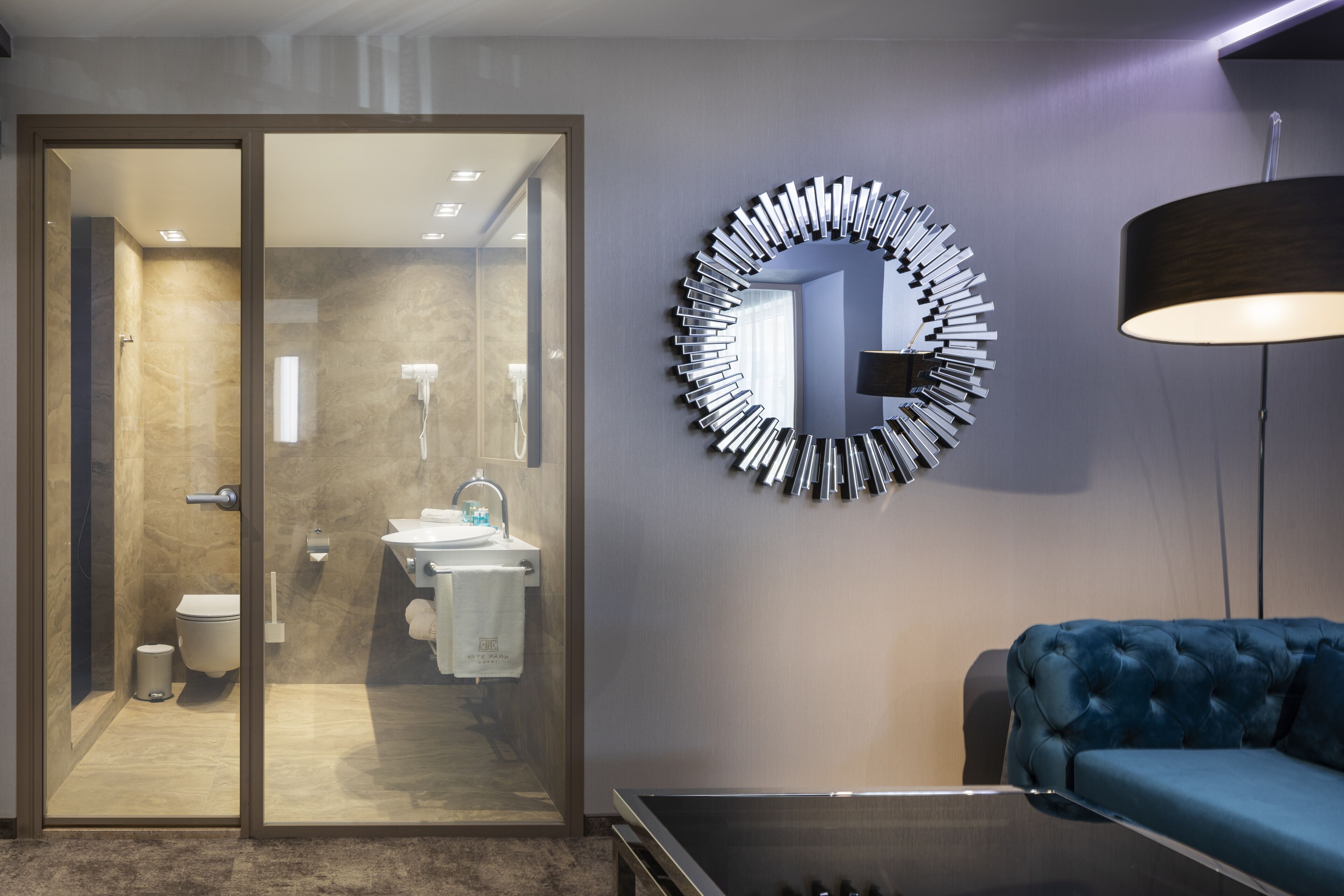 A modern bathroom with a glass shower enclosure, a round mirror, and a blue sofa in a living area.
