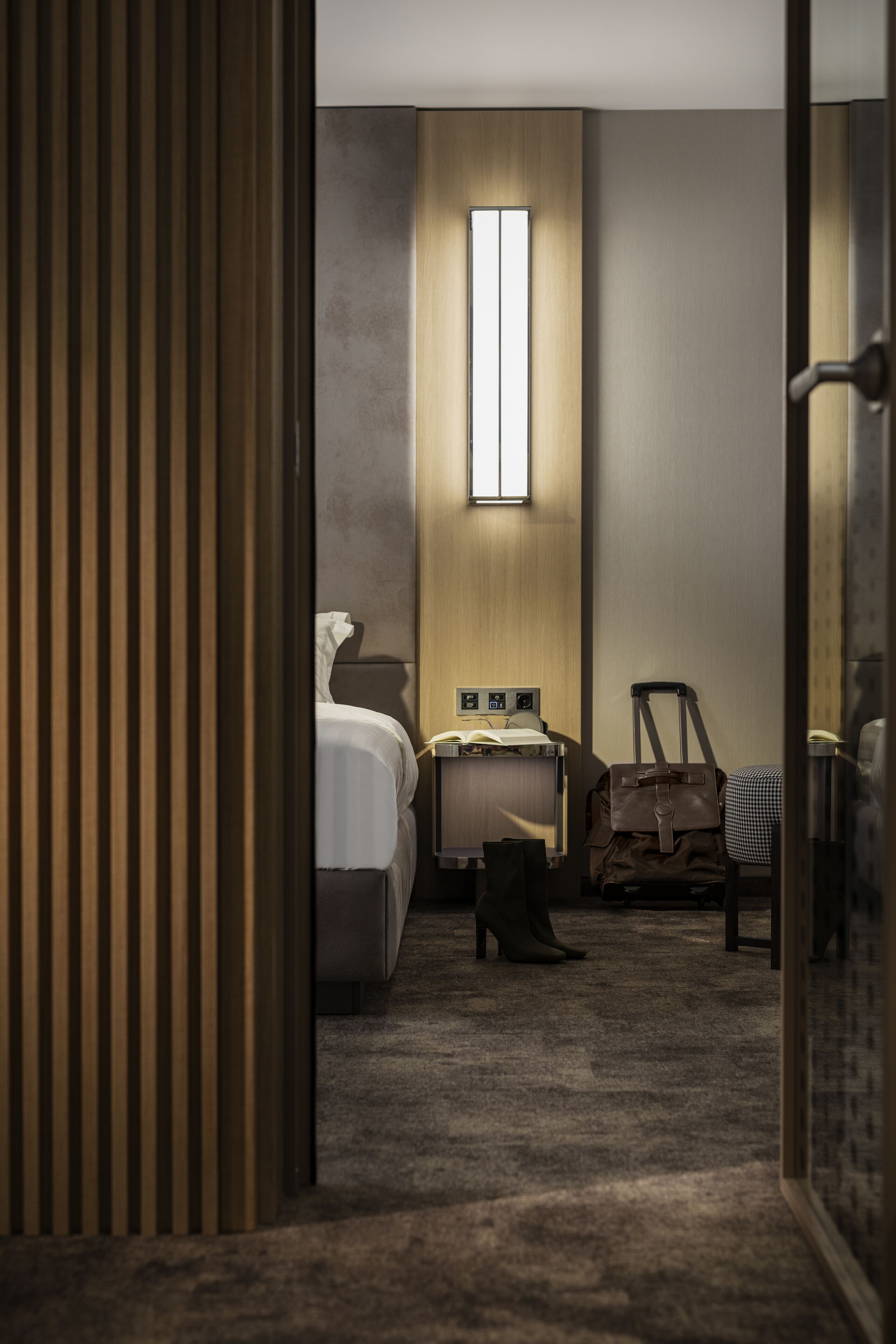 Habitación de hotel con cama, escritorio con lámpara, silla, maleta y ventana con luz natural.