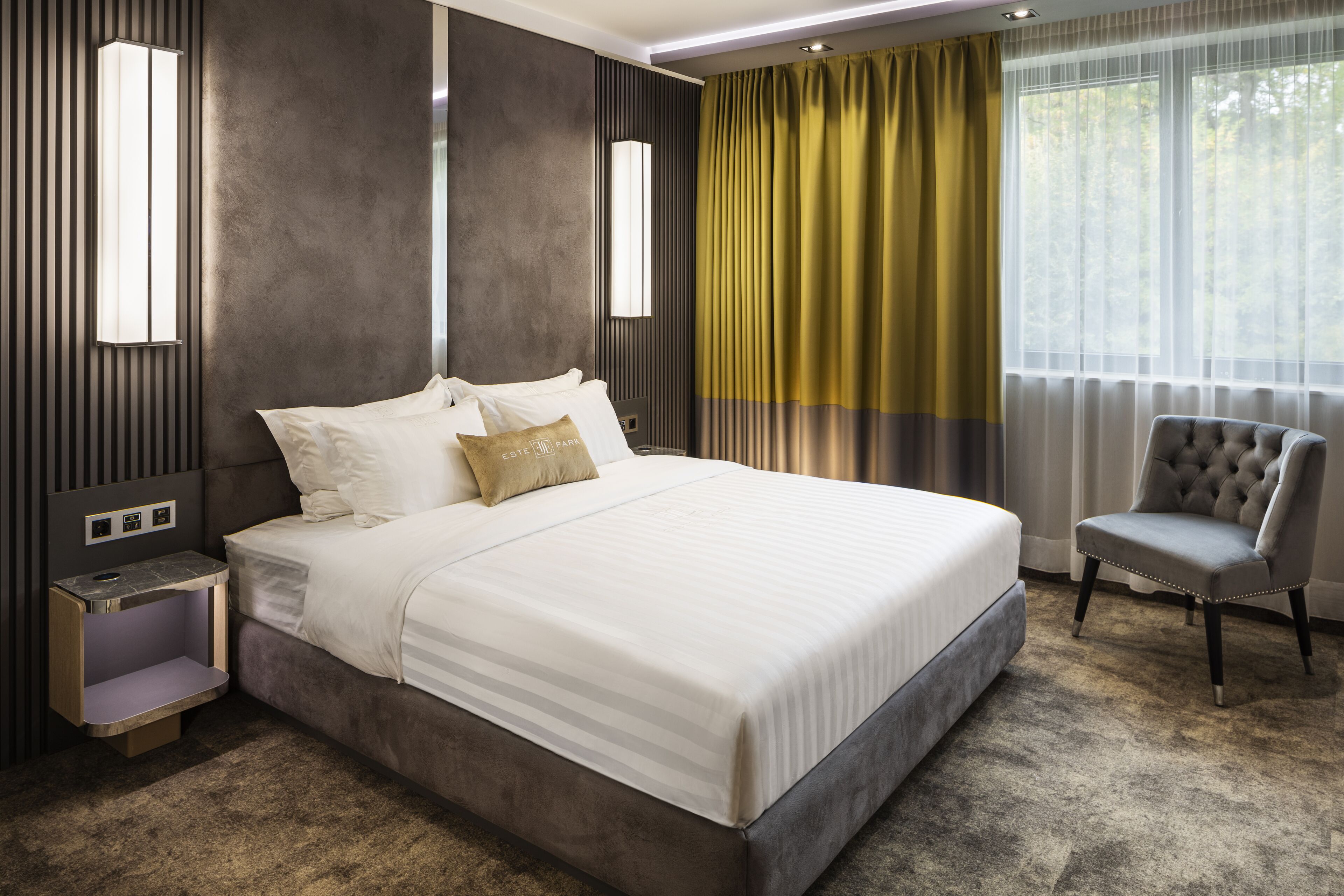 A modern hotel room with a large bed, a nightstand, a chair, and a window with curtains.
