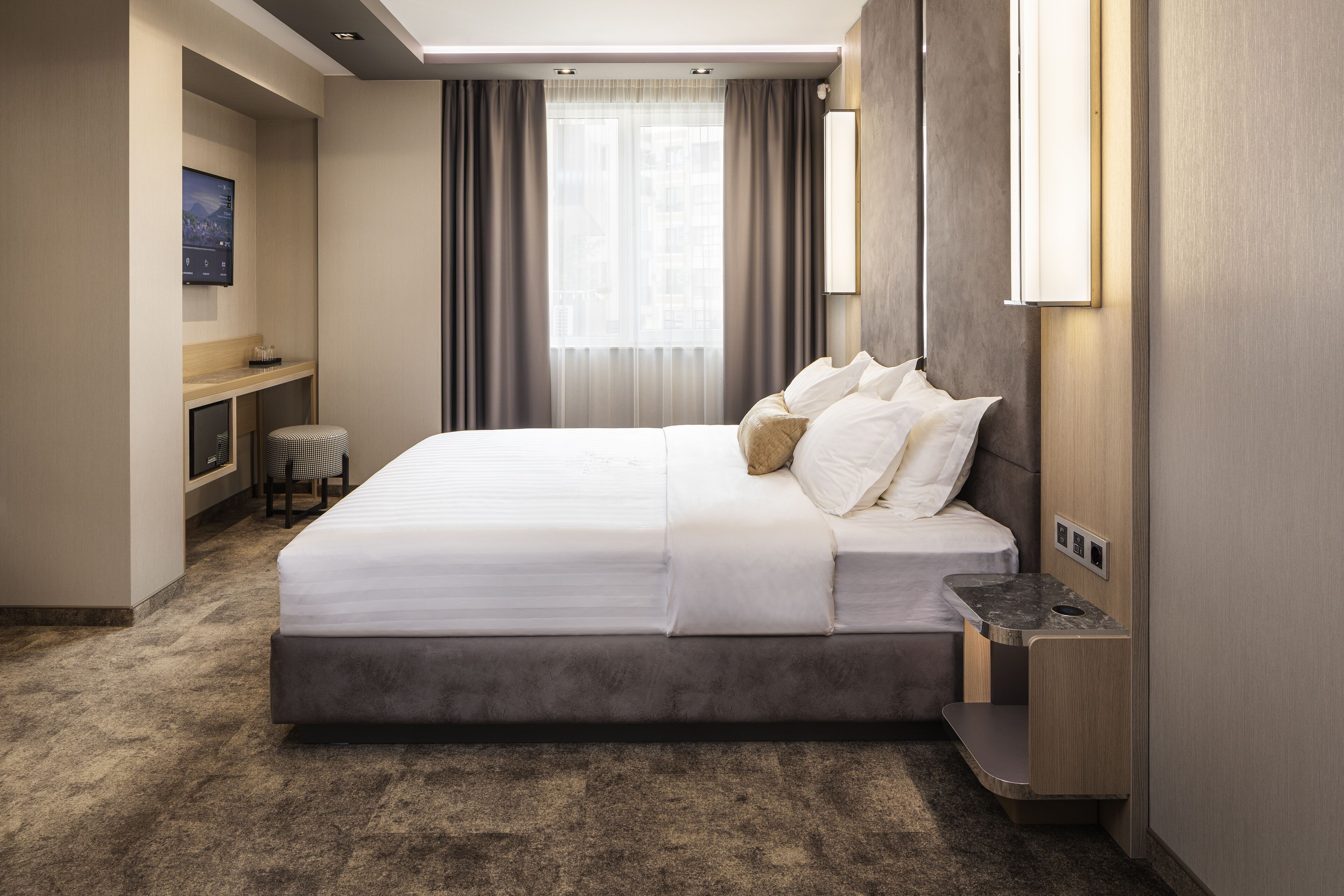 A modern hotel room with a large bed, a small desk, and a view of the city through the window.