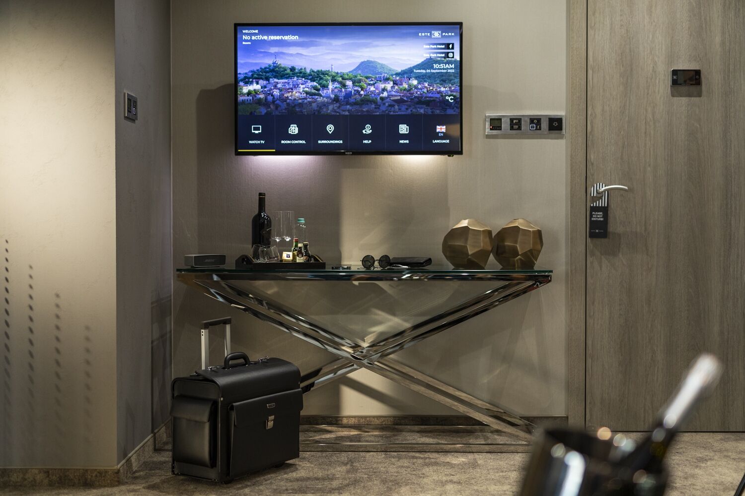 A modern hotel lobby with a TV displaying a welcome screen, a glass table with a bottle and glasses, and a suitcase on the floor.