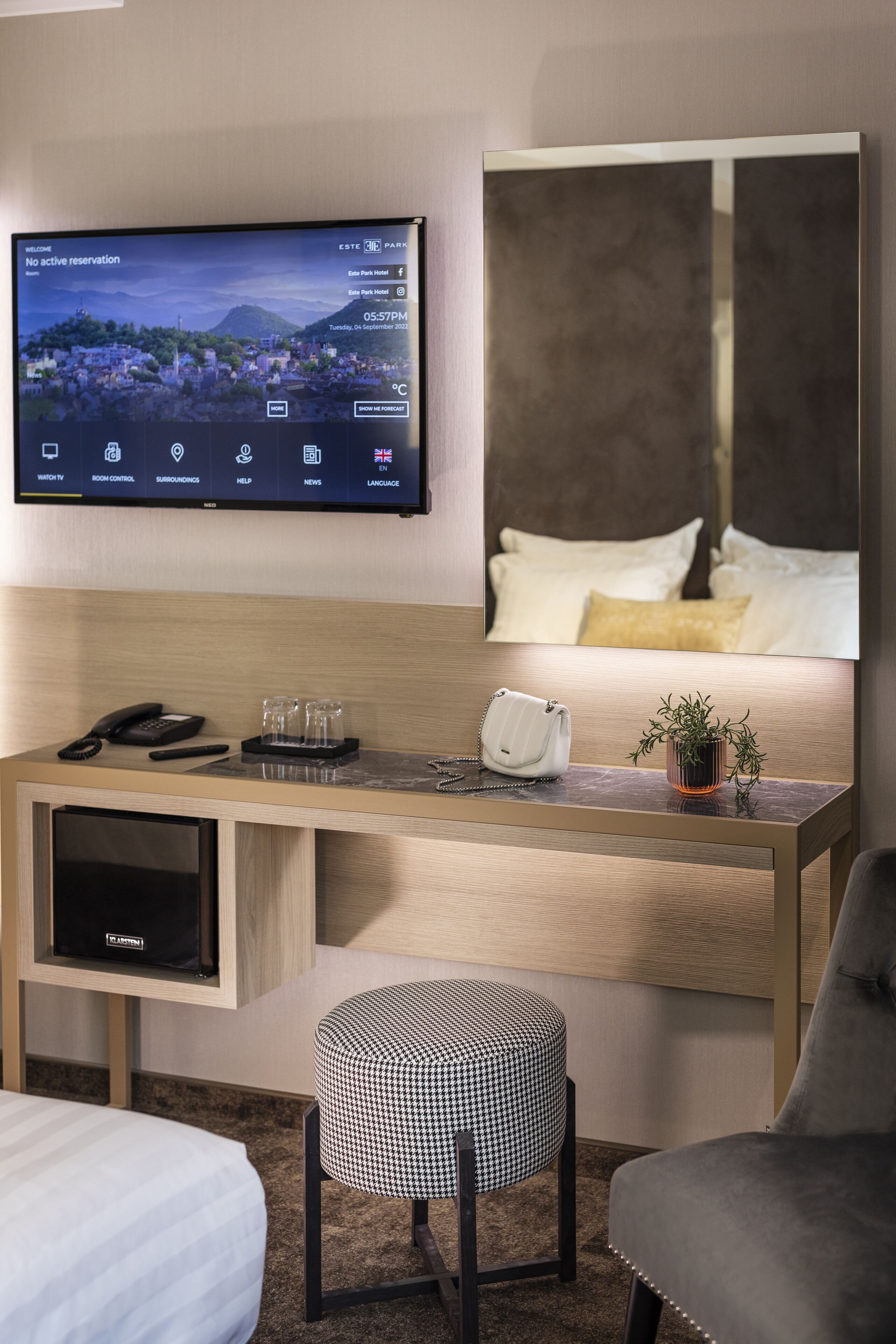 A hotel room with a flat-screen TV displaying a cityscape, a wooden desk with a phone, glasses, a handbag, and a plant.