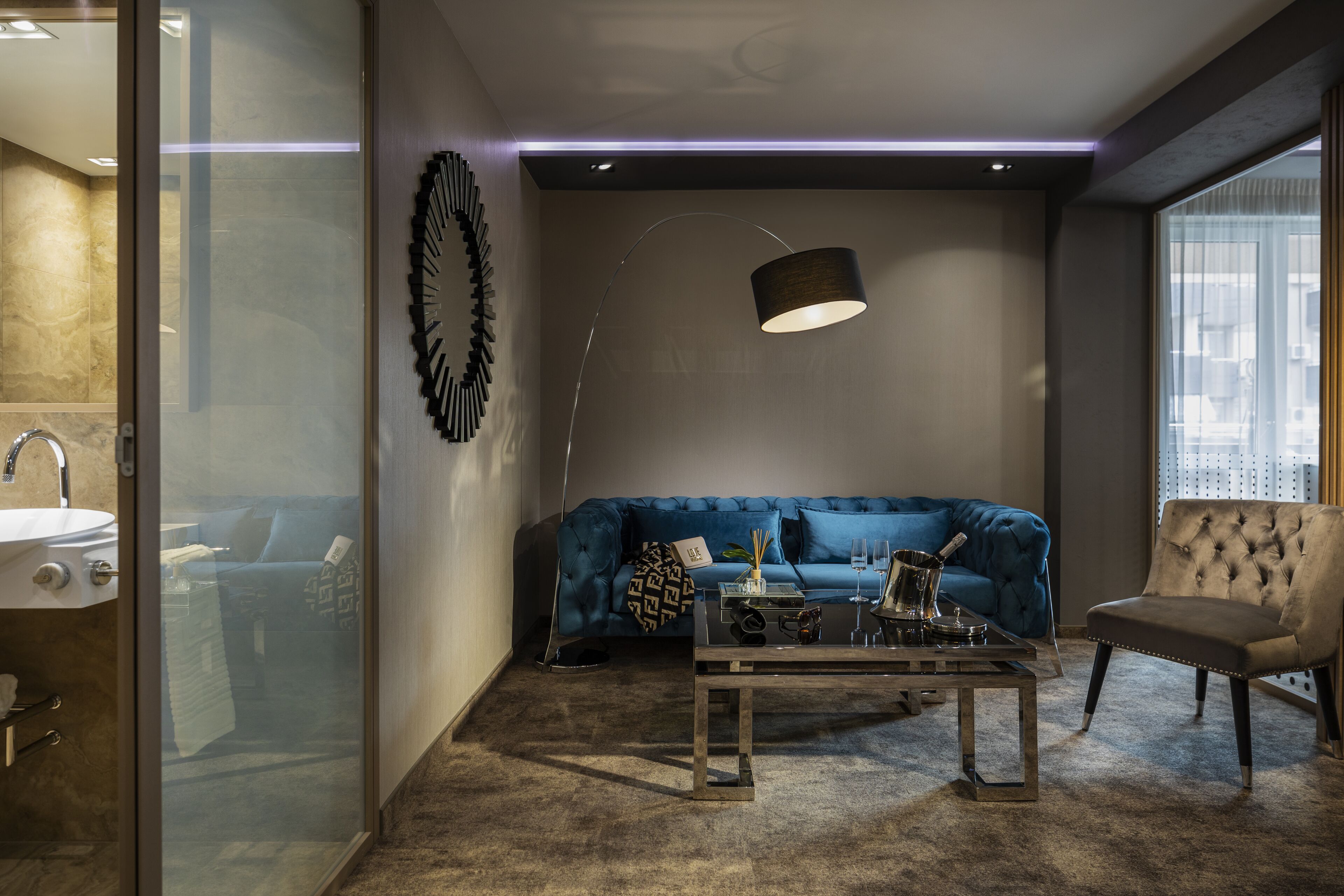 Un salon moderne avec un canapé bleu, un fauteuil capitonné, une table basse en verre et un lampadaire.